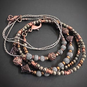 May include: A multi-strand bracelet featuring metallic copper-toned beads, dark grey porous stone beads, and small faceted metallic beads. The design includes ornate copper-colored spherical spacers and dark flower-shaped charms with a lobster clasp.