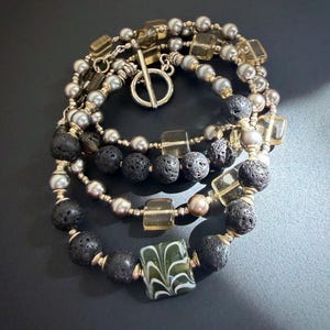 May include: A multi-strand bracelet featuring grey pearls, black porous lava beads, translucent square glass beads, and a single rectangular green glass bead with white swirl patterns. The piece is secured with a silver-toned toggle clasp.