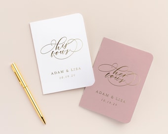 Puede incluir: Dos pequeños libros blancos y rosados con letras doradas. El libro blanco dice "His Vows" y el libro rosa dice "Her Vows". Ambos libros tienen los nombres "Adam & Lisa" y la fecha "10.19.20" impresa en ellos.