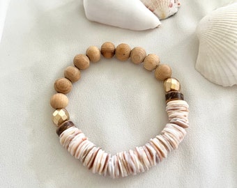 Può includere: Un braccialetto con perline di legno naturale, perline color oro e perline a forma di conchiglia. Il braccialetto è esposto su una superficie bianca con conchiglie sullo sfondo. Il braccialetto è di colore neutro. Le perline misurano circa 0,8 cm di diametro.