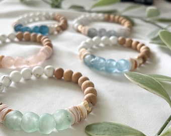 Può includere: Diversi braccialetti di perline con perline di legno naturale, perline bianche e varie pietre preziose, tra cui pietre blu e rosa. I braccialetti sono disposti su una superficie bianca, con bottiglie di oli essenziali e verde sullo sfondo.