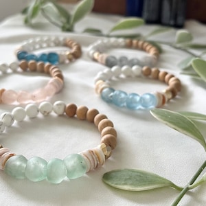 May include: Several beaded bracelets with natural wood beads, white beads, and various gemstones, including blue and pink stones. The bracelets are arranged on a white surface, with essential oil bottles and greenery in the background.
