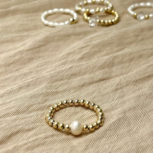 May include: A gold ring with a single pearl bead. The ring is on a brown fabric background.