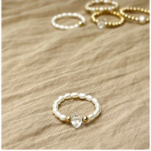 May include: A white pearl ring with a clear crystal heart charm. The ring is on a beige fabric background.