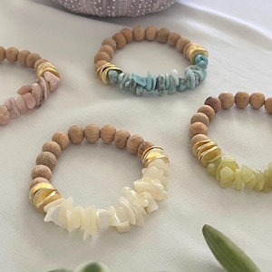 May include: Four beaded bracelets with natural wood beads and gold accents. Each bracelet features a different stone: pink, blue, white, and green. A purple sea urchin shell is in the background. The bracelets are on a white surface.
