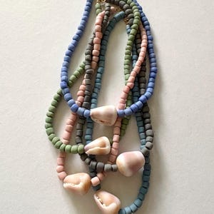 May include: A collection of handmade necklaces featuring colorful, square-shaped beads in blue, green, pink, and gray. Each necklace is adorned with a natural seashell pendant. The necklaces are connected with gold-colored clasps, creating a layered effect.