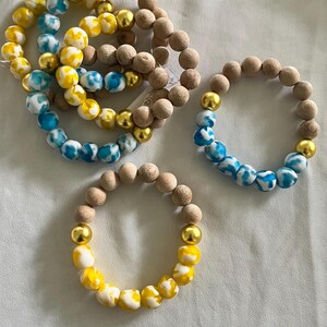 Puede incluir: Varios brazaletes de cuentas en una variedad de colores. Algunos brazaletes presentan cuentas de madera de color marrón claro, mientras que otros tienen cuentas estampadas azules, blancas y amarillas. Cuentas doradas acentúan algunos de los brazaletes.