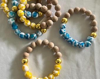 Può includere: Diversi braccialetti di perline in una varietà di colori. Alcuni braccialetti presentano perline di legno marrone chiaro, mentre altri hanno perline a fantasia blu, bianche e gialle. Perline color oro accentuano alcuni dei braccialetti.