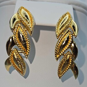 May include: A pair of gold-toned earrings with a leaf-like design. Each earring features three textured, elongated leaf shapes connected by curved, polished gold elements. The leaves have a detailed, textured surface with a darker center.