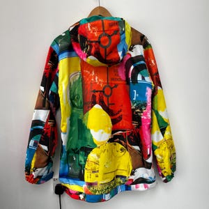 May include: A hooded jacket with a vibrant, abstract design. The jacket features a collage of colorful shapes, text, and patterns in red, yellow, green, and blue. The jacket is hanging on a wooden hanger.