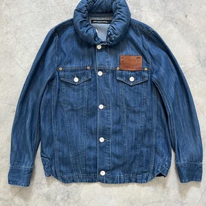 May include: A blue denim jacket with a distinctive, padded collar and white buttons. The jacket has two chest pockets and a brown leather patch. The garment is a medium wash denim, offering a classic and versatile style.