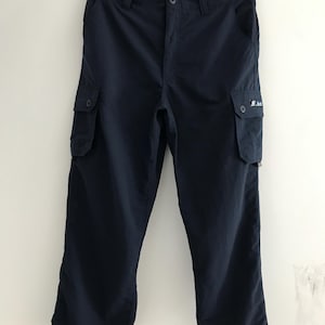 May include: Navy blue cargo pants hanging on a wooden hanger. The pants have two cargo pockets on the legs and a button closure. A small white logo is visible on one of the pockets. The pants are made of a lightweight material.