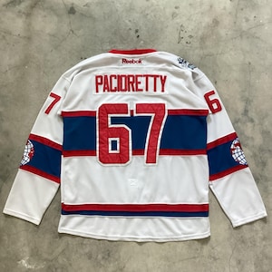 May include: White hockey jersey with red and blue accents. The jersey features the name "PACIORETTY" above the large number "67" in red. The number "7" is on the left sleeve and "6" on the right sleeve. The Reebok logo is visible at the top.