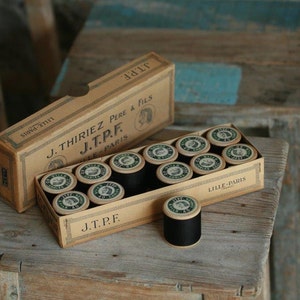 Puede incluir: Una caja de carretes de hilo negro con la etiqueta "J. Thiriez Pere & Fils J.T.P.F. Lille-Paris" en la parte delantera. La caja est&aacute; hecha de cart&oacute;n y tiene un aspecto vintage.