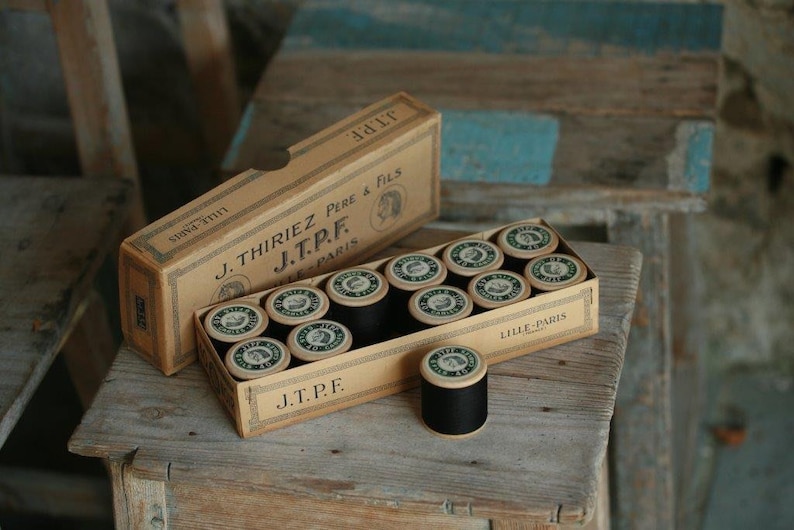 Puede incluir: Una caja de carretes de hilo negro con la etiqueta "J. Thiriez Pere & Fils J.T.P.F. Lille-Paris" en la parte delantera. La caja est&aacute; hecha de cart&oacute;n y tiene un aspecto vintage.