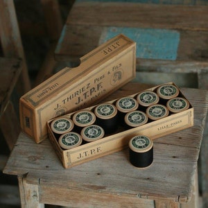 Puede incluir: Una caja de carretes de hilo negro con la etiqueta "J. Thiriez Pere & Fils J.T.P.F. Lille Paris" en la caja.