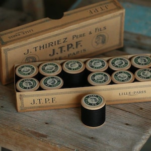 Puede incluir: Una caja de carretes de hilo negro con etiquetas verdes y blancas. La caja es marr&oacute;n y tiene el texto "J.Thiriez Pere & Fils J.T.P.F. Lille-Paris" impreso en ella.