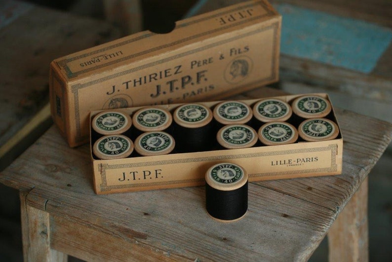 Puede incluir: Una caja de carretes de hilo negro con etiquetas verdes y blancas. La caja es marr&oacute;n y tiene el texto "J.Thiriez Pere & Fils J.T.P.F. Lille-Paris" impreso en ella.