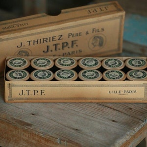 Puede incluir: Una caja vintage de carretes de hilo etiquetados "J.Thiriez Pere & Fils J.T.P.F. Lille-Paris (Francia)". La caja es de cart&oacute;n y tiene un esquema de color marr&oacute;n y blanco. Los carretes son de metal y tienen una etiqueta verde y blanca.