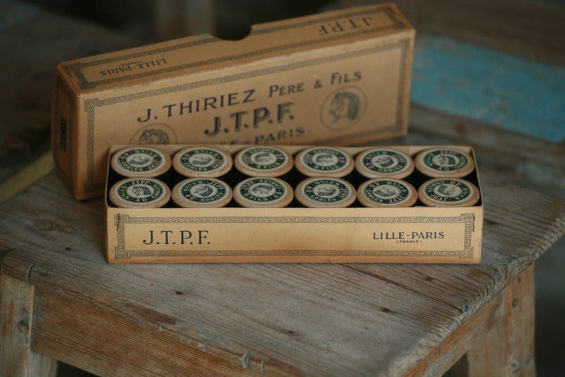 Puede incluir: Una caja vintage de carretes de hilo etiquetados "J.Thiriez Pere & Fils J.T.P.F. Lille-Paris (Francia)". La caja es de cart&oacute;n y tiene un esquema de color marr&oacute;n y blanco. Los carretes son de metal y tienen una etiqueta verde y blanca.