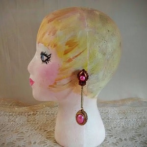 May include: A mannequin head with painted blonde hair and pink and gold dangle earrings. The earrings have a pink pearl and a gold filigree design.