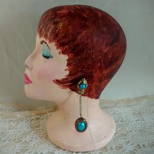May include: A painted plaster mannequin head with short, auburn hair and a turquoise and gold dangle earring.
