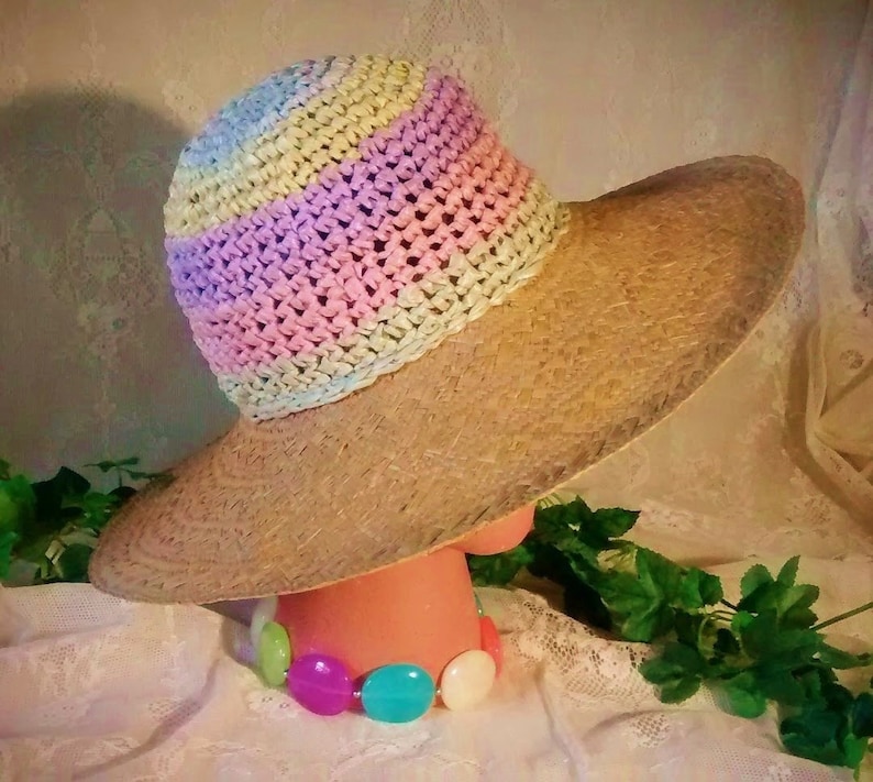 Puede incluir: Un sombrero de paja de ala ancha con una corona de crochet en rayas de color rosa pastel, amarillo y azul. El sombrero est&aacute; sobre una superficie blanca con un collar de cuentas de colores.