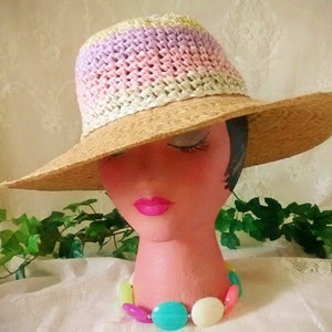 Puede incluir: Un sombrero de paja de ala ancha con una banda de crochet en tonos de rosa, morado y blanco. El sombrero est&aacute; sentado en una cabeza de maniqu&iacute; con un collar de cuentas de colores.