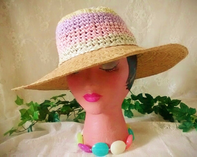 Puede incluir: Un sombrero de paja de ala ancha con una banda de crochet en tonos de rosa, morado y blanco. El sombrero est&aacute; sentado en una cabeza de maniqu&iacute; con un collar de cuentas de colores.