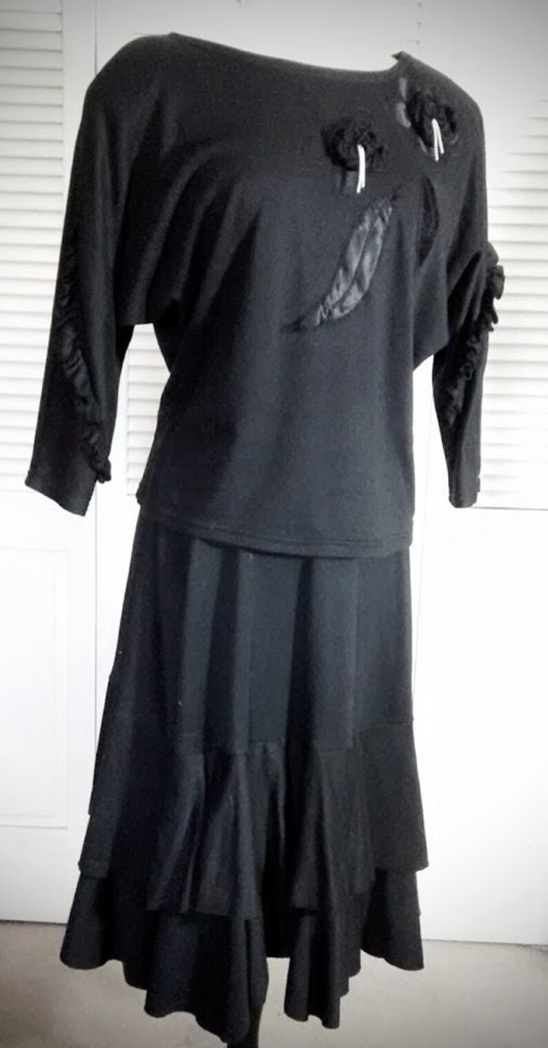 May include: A black two-piece outfit with a ruffled skirt and a long-sleeved top with black fabric flowers and a black feather detail.