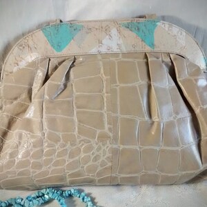 May include: A beige crocodile-embossed handbag with a flap closure and a decorative top made of white and turquoise fabric.