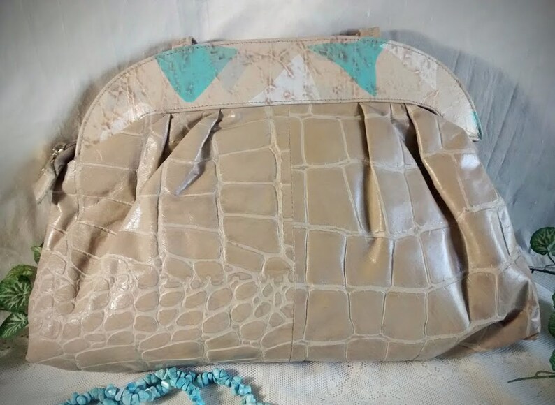 May include: A beige crocodile-embossed handbag with a flap closure and a decorative top made of white and turquoise fabric.