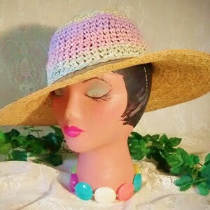 Puede incluir: Un sombrero de paja de ala ancha con una corona de ganchillo en tonos de rosa, azul y blanco. El sombrero est&aacute; sentado en una cabeza de maniqu&iacute; con un collar de cuentas de colores.