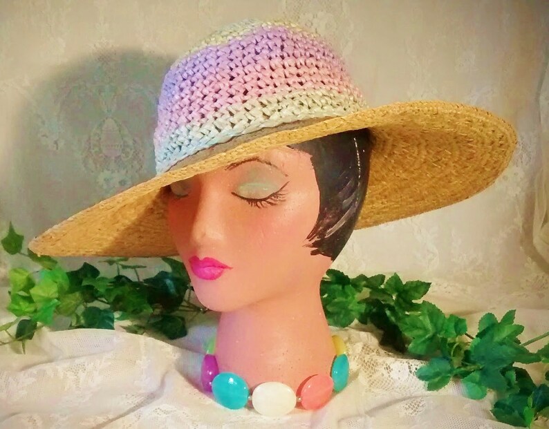 Puede incluir: Un sombrero de paja de ala ancha con una corona de ganchillo en tonos de rosa, azul y blanco. El sombrero est&aacute; sentado en una cabeza de maniqu&iacute; con un collar de cuentas de colores.