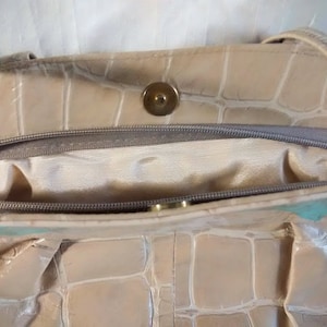 May include: A beige leather handbag with a crocodile embossed pattern. The bag has a zipper closure and a gold-toned button.