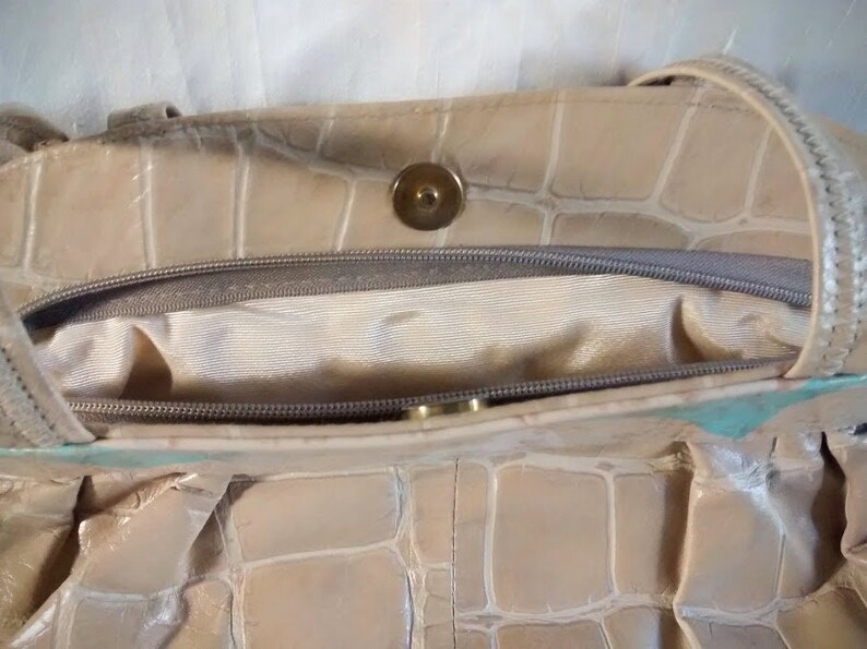 May include: A beige leather handbag with a crocodile embossed pattern. The bag has a zipper closure and a gold-toned button.