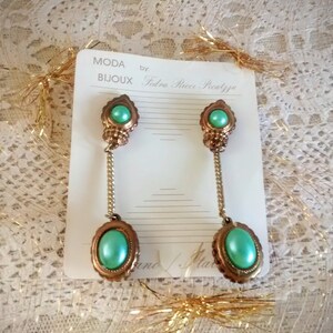 May include: A pair of long, dangling earrings with green stones and gold accents. The earrings are on a white card with the text "Moda Bijoux by Fedora Ricci Piccaluga".