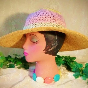 Puede incluir: Un sombrero de paja de ala ancha con una corona de ganchillo en tonos de rosa, blanco y verde. El sombrero est&aacute; sentado en una cabeza de maniqu&iacute; con un collar de cuentas de colores.