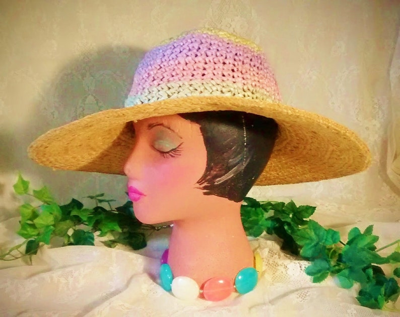 Puede incluir: Un sombrero de paja de ala ancha con una corona de ganchillo en tonos de rosa, blanco y verde. El sombrero est&aacute; sentado en una cabeza de maniqu&iacute; con un collar de cuentas de colores.