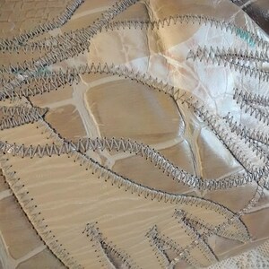 May include: Close-up of a beige leather bag with a unique patchwork design. The bag is made of several pieces of leather sewn together with a decorative stitch. The leather has a textured, crocodile-like pattern.