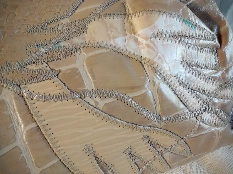 May include: Close-up of a beige leather bag with a unique patchwork design. The bag is made of several pieces of leather sewn together with a decorative stitch. The leather has a textured, crocodile-like pattern.