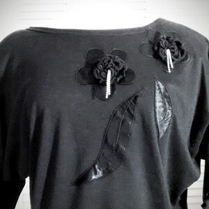 May include: Black long-sleeved shirt with a scoop neck and decorative black fabric flowers and leaves.