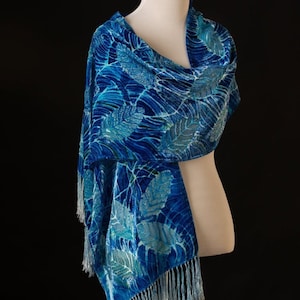 Puede incluir: Una bufanda de seda azul con un patrón de hojas blancas y flecos. La bufanda está drapeada sobre un maniquí blanco.