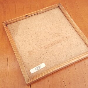 May include: A square wooden picture frame with a light brown finish. The frame has a natural wood grain and a light-coloured backing. A small white sticker with text is visible on the back. The frame is on a wooden surface.