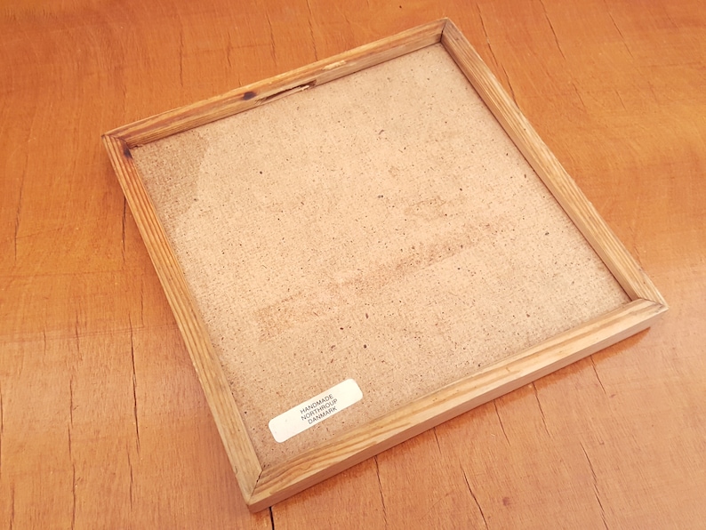 May include: A square wooden picture frame with a light brown finish. The frame has a natural wood grain and a light-coloured backing. A small white sticker with text is visible on the back. The frame is on a wooden surface.