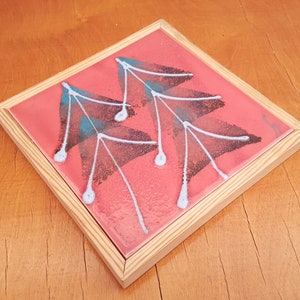 May include: Square decorative tile with a pink background and a leaf design in black, teal, and white. The tile is set in a natural wood frame. The artwork is a unique piece of home decor.