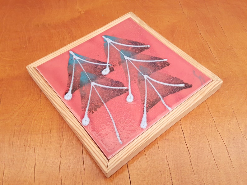 May include: Square decorative tile with a pink background and a leaf design in black, teal, and white. The tile is set in a natural wood frame. The artwork is a unique piece of home decor.