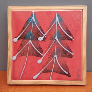 May include: Square ceramic tile with a red background and four abstract tree designs in black, teal, and white. The tile is set in a natural wood frame, creating a decorative piece for wall art or home decor.
