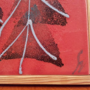 May include: A framed ceramic tile with a red background and an abstract design. The design features black and white shapes with white lines radiating outwards. The tile is set in a natural wood frame. A signature is visible in the bottom right corner.