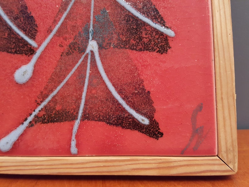 May include: A framed ceramic tile with a red background and an abstract design. The design features black and white shapes with white lines radiating outwards. The tile is set in a natural wood frame. A signature is visible in the bottom right corner.